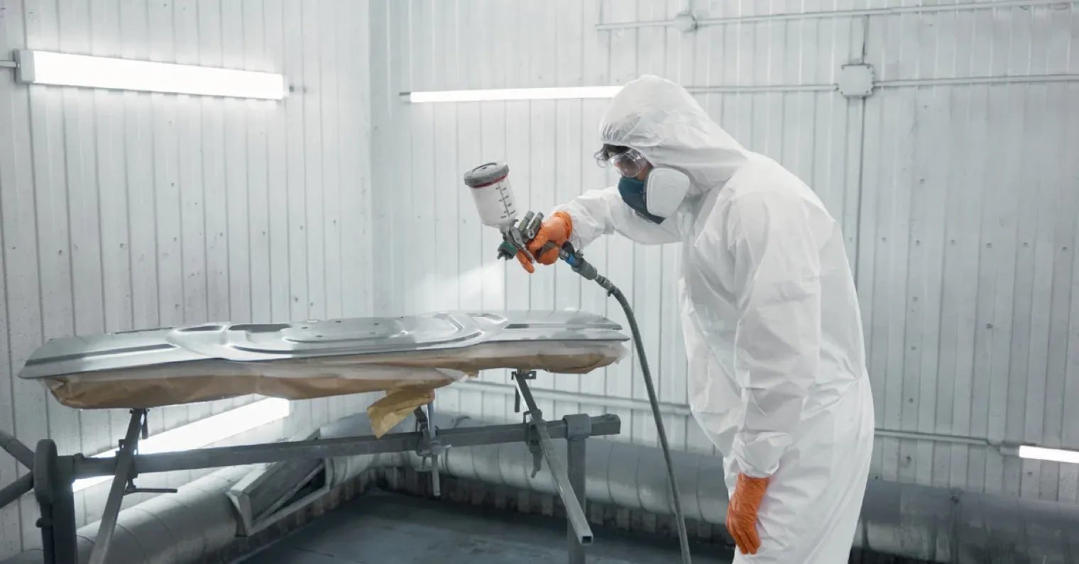 A painter in a respirator hand-spraying a panel inside a traditional spray booth